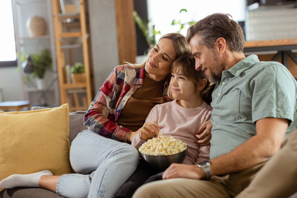 Uma família de três pessoas está confortavelmente aninhada em um sofá, seus rostos refletindo emoção e atenção enquanto compartilham uma tigela de pipocas durante uma noite de suspense - Foto, Imagem