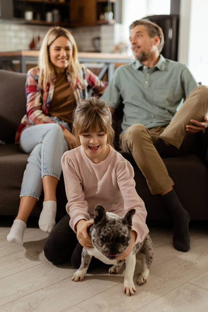 Fröhliche Eltern sehen zu, wie ihre Tochter zu Hause mit einer fröhlichen französischen Bulldogge spielt - Foto, Bild