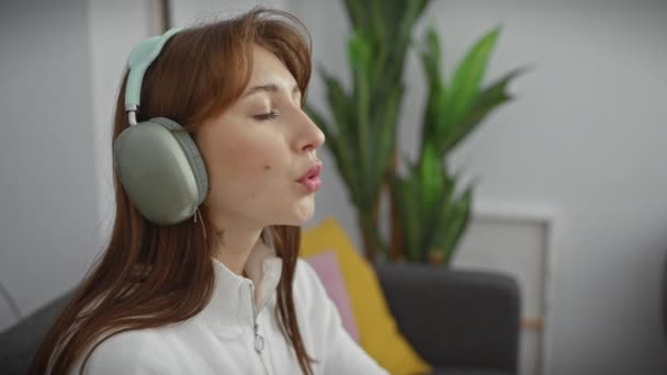 Kulaklıklı genç bir kadın evde müzik dinliyor, modern bir oturma odasında eğlence ve teknoloji sergiliyor.. - Video, Çekim