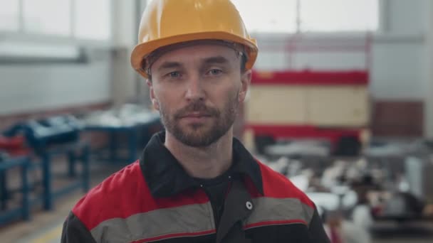 Valkoihoisen teollisuustyöntekijän rinta yllään keltainen kypärä ja työvaatteet katsoen kameraa, joka seisoo hämärtyneellä taustalla sisätiloissa tehtaalla - Materiaali, video