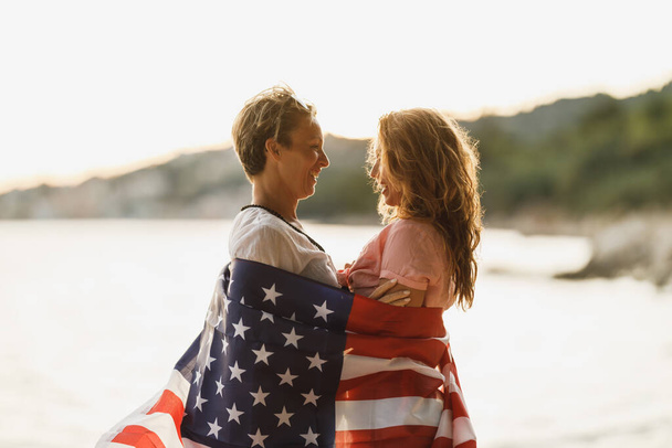 ビーチでリラックスした一日を楽しむ米国国旗を持つ2人の愛する女性の友人. - 写真・画像