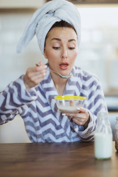 Mujer joven en albornoz comiendo avena instantánea para el desayuno en la cocina en casa. - Foto, imagen