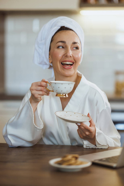 Μια ελκυστική νεαρή γυναίκα με μπουρνούζι χαλαρώνει και πίνει πρωινό καφέ στο σπίτι της.. - Φωτογραφία, εικόνα