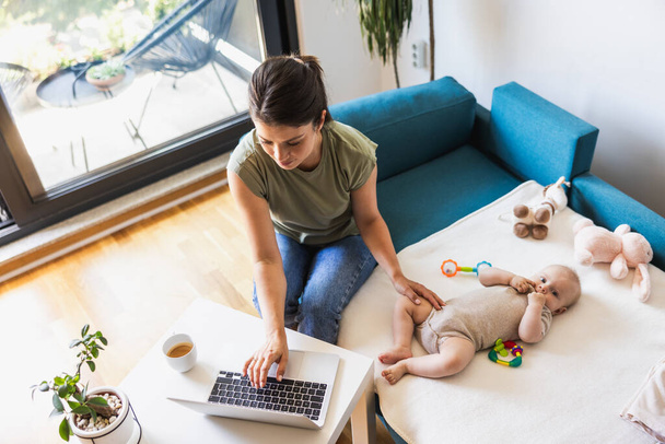 Evde dizüstü bilgisayarla çalışırken bebeğine bakan genç bir annenin fotoğrafı.. - Fotoğraf, Görsel