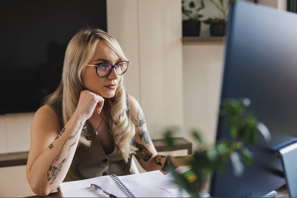 Joven diseñadora freelancer femenina que se ve pensativa mientras trabaja en la computadora desde su oficina en casa. - Foto, imagen
