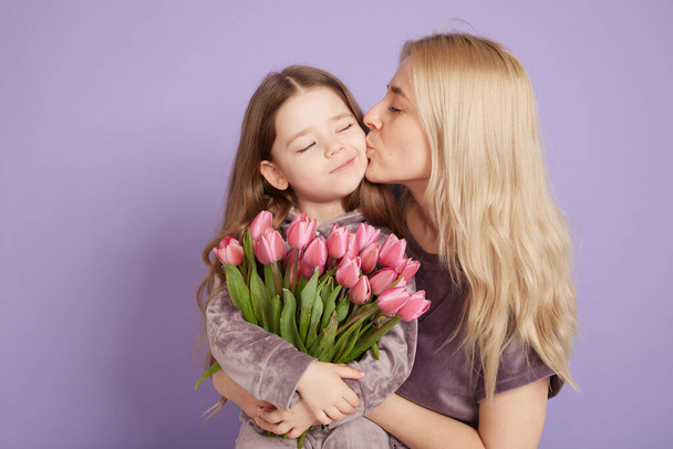 Ein kleines Mädchen mit seiner Mutter und einem Blumenstrauß umarmen sich auf lila Hintergrund, Familienurlaub und Zweisamkeit. - Foto, Bild