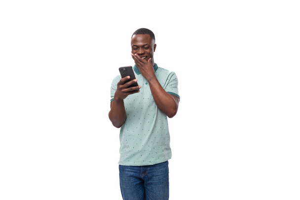 νεαρός όμορφος Αφρικανός με κοντό κούρεμα ντυμένος με μπλουζάκι και τζιν μέντας επικοινωνεί στα κοινωνικά δίκτυα σε ένα smartphone. - Φωτογραφία, εικόνα