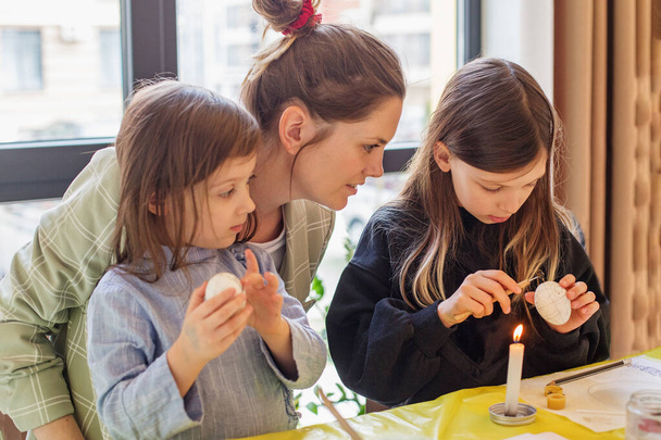 Tierno momento familiar como madre enseña a sus dos hijas el arte de la decoración de huevos de Pascua en su casa. - Foto, Imagen