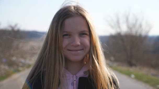 Close up of happy little girl looks into camera outdoor and laughs covering her face with hand. Portrait of funny female child with long blonde hair stands on road at countryside with joyful smile. - Filmagem, Vídeo