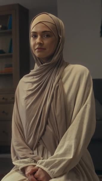 Střední vertikální portrét atraktivní mladé muslimky v světle béžové hidžáb a abája pózující sedí na kolenou na podlaze v majlis a dívá se na kameru - Záběry, video