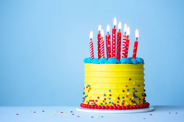 Sarı kremalı renkli doğum günü pastası ve mavi arka planda kırmızı doğum günü mumları. - Fotoğraf, Görsel
