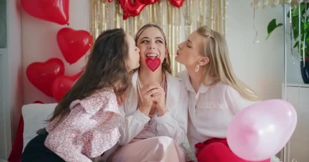 Alegres mujeres jóvenes besando a una amiga sosteniendo caramelos en forma de corazón durante la celebración del día de Galentines en casa - Metraje, vídeo