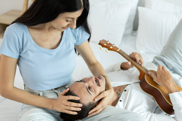 幸せなカップルはベッドでウクレレを演奏する. 音楽を楽しんでいる男性と女性は,家族の中で幸せな活動を生み出します. 楽器とロマンチックな若いカップル楽しい幸せ. - 写真・画像