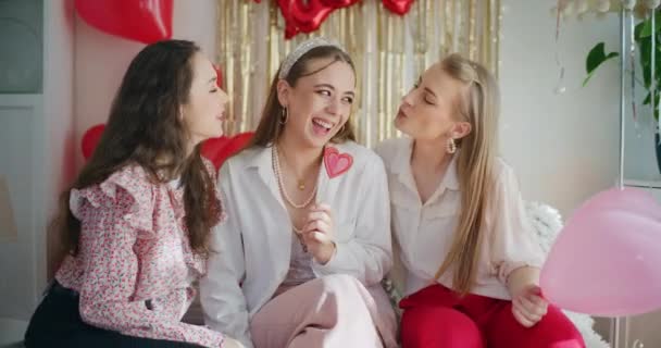 Счастливые молодые женщины целуются друг с другом держа конфеты в форме сердца во время празднования дня Галинцев дома - Кадры, видео