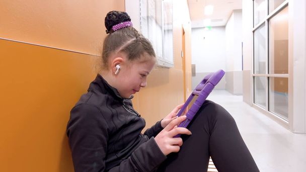 Genesteld in een rustige gang, vindt deze toegewijde jonge kunstschaatser een moment van rust, verdiept in haar tablet terwijl ze zich mentaal voorbereidt op haar komende optreden.. - Foto, afbeelding