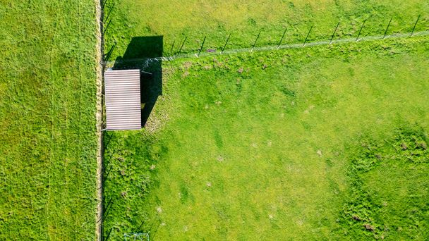 Esta fotografía aérea captura un pequeño cobertizo solitario con un techo a dos aguas en medio de un pasto verde vibrante. La estructura proyecta una sombra nítida en el suelo, lo que indica que el sol está en su cenit. - Foto, Imagen