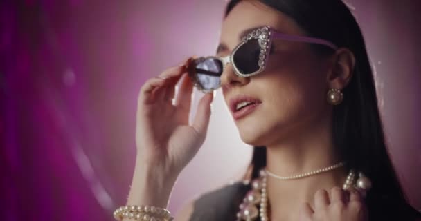 Γοητευτική νεαρή γυναίκα σε μοντέρνα γυαλιά ηλίου και μαργαριτάρι κοσμήματα μπροστά από φωτισμένο φόντο - Πλάνα, βίντεο
