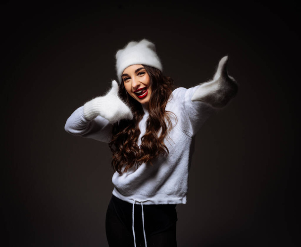 A woman in a white sweater and white hat is smiling and giving a thumbs up. The image has a happy and positive mood - Foto, Imagen