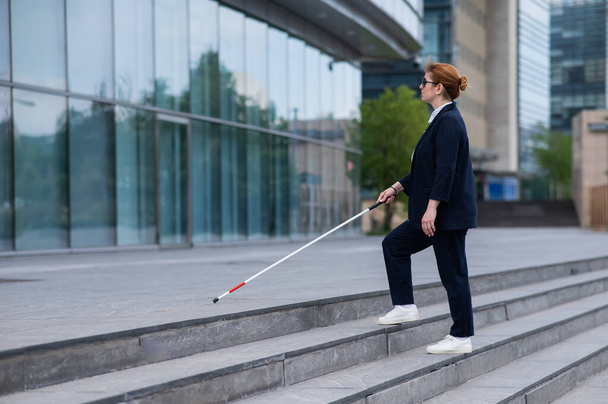 Blind business woman in glasses and with a cane climbs the stairs to the business center - 写真・画像