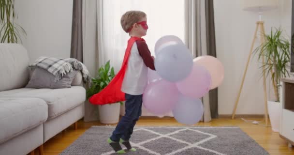 Een jong kind in een superheld kostuum speelt met kleurrijke ballonnen in een gezellige woonkamer. Hoge kwaliteit 4k beeldmateriaal - Video