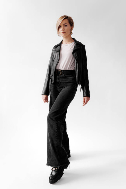 Una giovane donna in bilico si trova con fiducia in una giacca di pelle e jeans svasati, incarnando urban chic - Foto, immagini