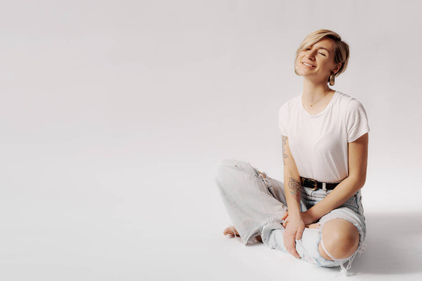 Una joven alegre sentada en una pose relajada, sonriendo con los ojos cerrados en un denim ligero y una camiseta blanca - Foto, imagen