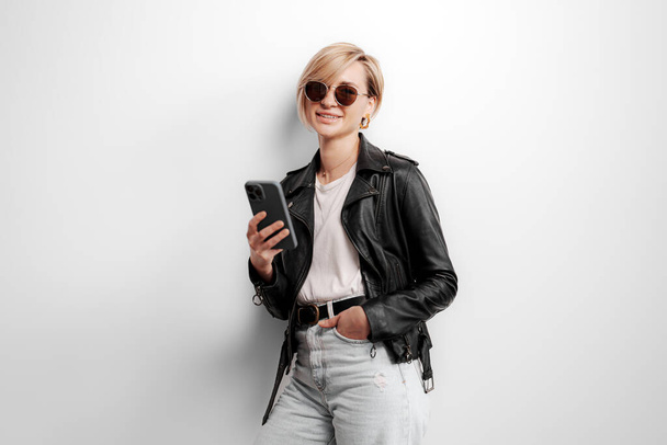 Модная молодая женщина взаимодействует со смартфоном, носит кожаную куртку и солнцезащитные очки для технически подкованного взгляда - Фото, изображение