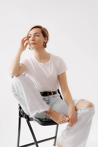 Споглядальна молода жінка позує в білому трійнику і легкому джинсі, сидячи на сучасному стільці, виключаючи випадкову елегантність - Фото, зображення