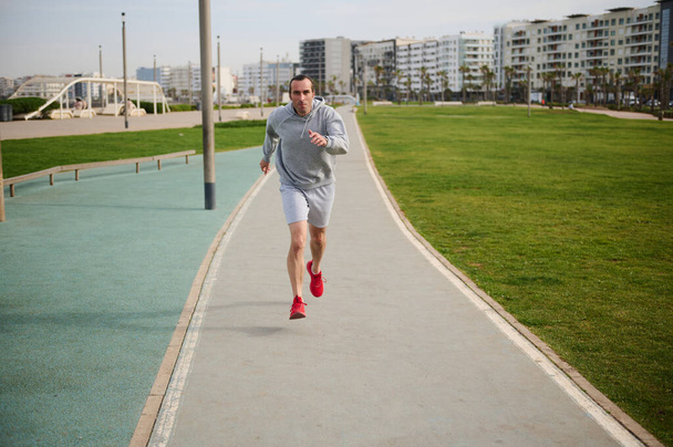Dlouhý záběr zdravého mladého muže v šedých sportovních šatech a červených teniskách, běžícího po promenádě. Samec běžec sprintující na běžeckém pásu venku. - Fotografie, Obrázek