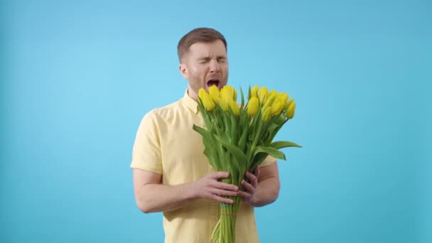 Hombre oliendo ramo de flores amarillas y estornudar sobre fondo azul - Imágenes, Vídeo