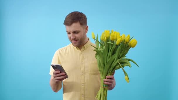 Hombre con teléfono y ramo de flores amarillas sobre fondo azul - Metraje, vídeo