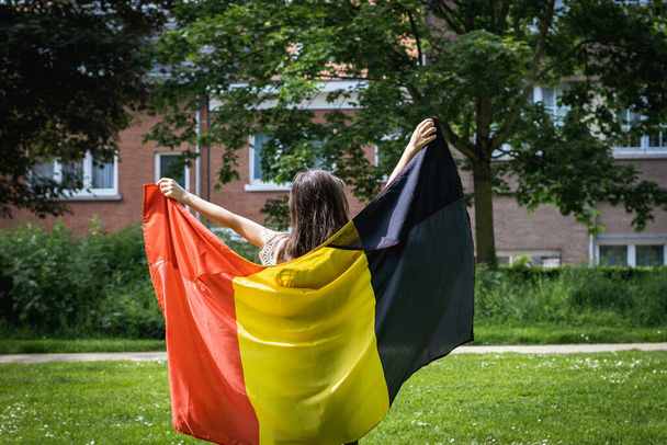 Портрет однієї красивої кавказької дівчинки брюнетки з вільним волоссям, що стоїть ззаду і тримає бельгійський прапор, відкритий в міському парку в літній день, вид крупним планом. - Фото, зображення