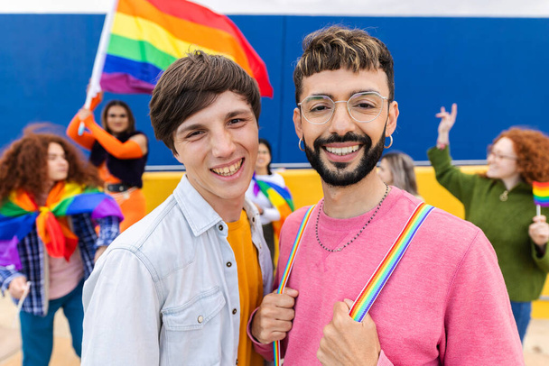 Retrato de dos hombres homosexuales activistas por los derechos LGBTI sonriendo a cámara sobre diversas personas homosexuales celebrando el día del orgullo gay en el fondo. Concepto de comunidad LGBT. - Foto, Imagen