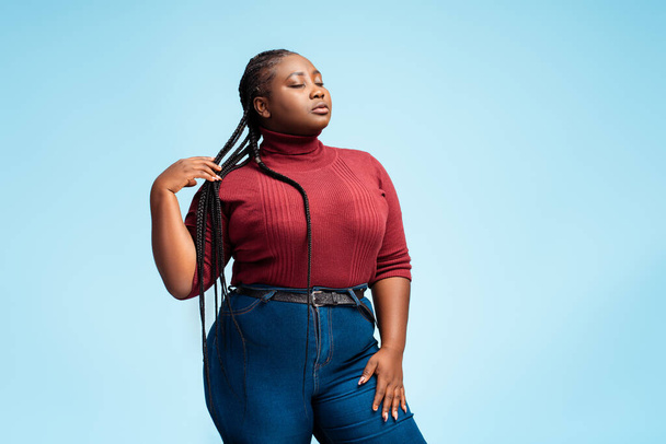 Pensive attractive African American woman with stylish braids, wearing casual clothing, red sweater and denim with closed eyes posing isolated on blue background. Hair care concept - Photo, Image