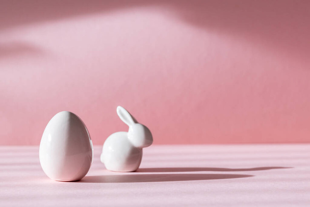 Figuras de porcelana de ovos de Páscoa e coelho ficam à esquerda em um fundo rosa com sombras e espaço de cópia à direita, vista lateral close-up. - Foto, Imagem