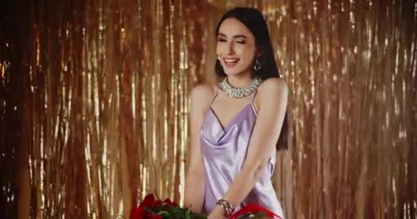 Sorrindo na moda atraente jovem segurando um monte de rosas vermelhas de pé contra a decoração brilhante streamer - Filmagem, Vídeo
