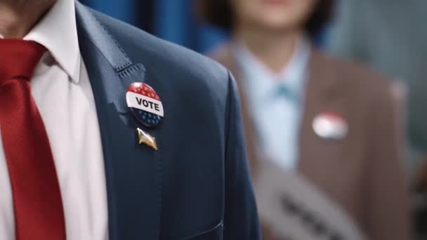 Close-up tiro de parte superior do corpo do candidato masculino irreconhecível em terno, camisa branca e gravata vermelha, com emblemas de bandeira Vote e EUA, dando discurso sincero com a mão no coração durante convenção eleitoral - Filmagem, Vídeo