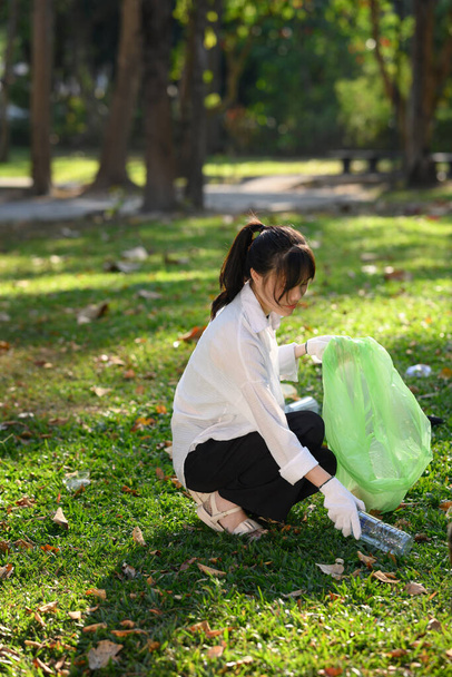Portrait of young woman picking up litter in the park on a sunny bright day. Environmental protection concept. - Zdjęcie, obraz