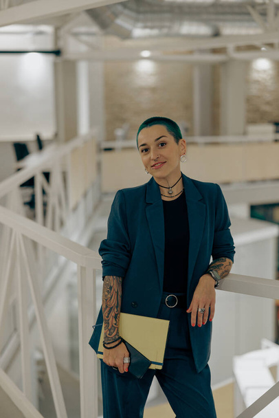Usmívající se potetovaná podnikatelka se zelenými krátkými vlasy držící notebook stojící v moderní kanceláři - Fotografie, Obrázek