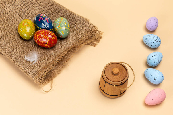Színes húsvéti tojások egy zsákvásznon, egy kis fahordó a bézs háttéren. Felülnézet. - Fotó, kép