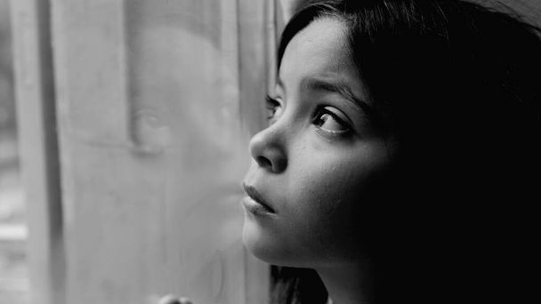 One unhappy somber little girl feeling depressed during hard times. Close-up child's face and hand leaning on glass window feeling lonely and solitude in black and white, monochromatic - Photo, Image