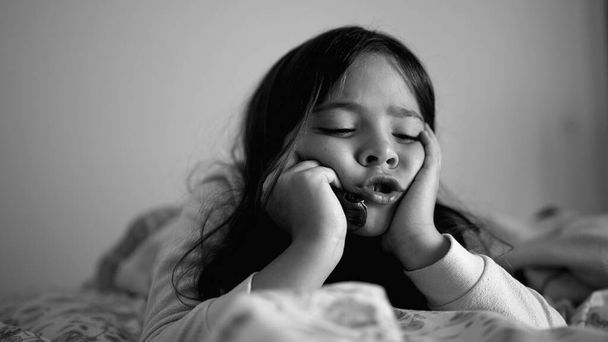 Sad depressed little girl laid in bed holding head with hands in chin, close-up face of melancholic child feeling down in black and white, monochrome - Photo, Image