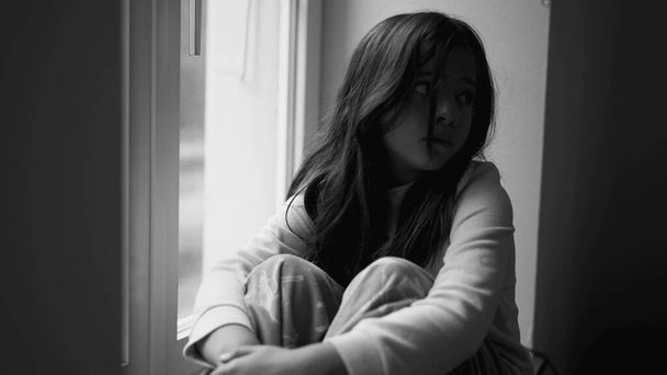 Депрессивная маленькая девочка глядя на вид из окна квартиры, потерял в мысли и чувство грусти в драматическом черно-белом, детские психические заболевания - Фото, изображение