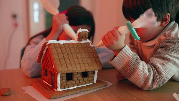 Familie binding over versieren peperkoek huis, kinderen zorgvuldig toepassen van Royal Icing, voorbereiding op vreugdevolle feestdagen - Foto, afbeelding