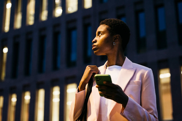若い黒人のスタイリッシュなビジネスマンは,彼女の手を離れて見ている携帯電話を持っています. 夜にダウンタウンのディクトリに携帯電話を持っているビジネスマン. - 写真・画像
