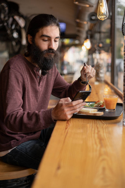 Hipster γενειοφόρος άνθρωπος τρώει το μεσημεριανό του κάθεται στο καφέ και να χρησιμοποιήσετε το κινητό τηλέφωνο για sms γραπτών μηνυμάτων, ανάγνωση ειδήσεων. Αντιγραφή χώρου - Φωτογραφία, εικόνα