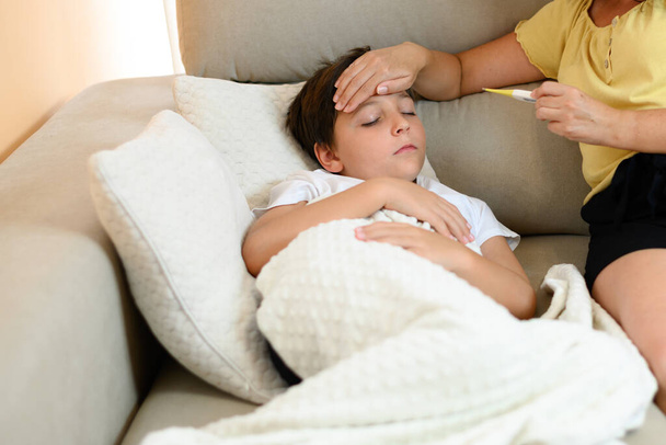Sick child on the couch at home. Someone puts their hand on the child's forehead and looks at the thermometer. - Photo, Image