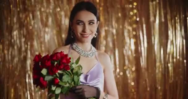 Smiling attractive young woman holding bunch of red roses while standing against shiny streamer decoration - Footage, Video
