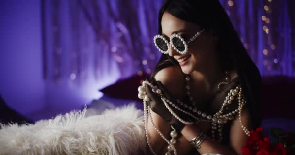 Χαμογελώντας ελκυστική γυναίκα σε μαργαριτάρια γυαλιά κρατώντας κοσμήματα, ενώ βρίσκεται στο κρεβάτι στο φωτεινό υπνοδωμάτιο - Πλάνα, βίντεο