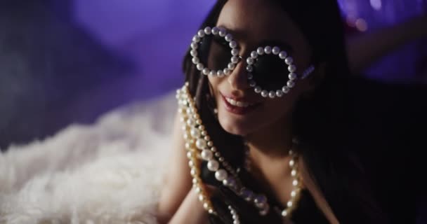 Detailní záběr na usmívající se trendy mladá žena v perleťových brýlích drží šperky při ležení na posteli - Záběry, video